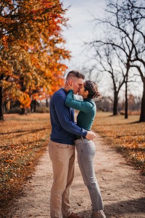 A couple embraces on a dirt path surrounded by autumn trees with vibrant orange foliage.