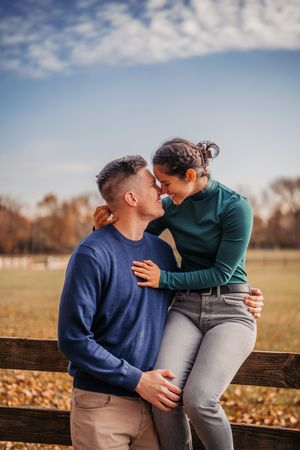 A couple shares an intimate moment while sitting on a wooden fence in an autumn field during golden hour.