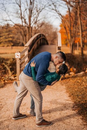 Two people share an intimate embrace in a park during autumn with fallen orange leaves on the ground.