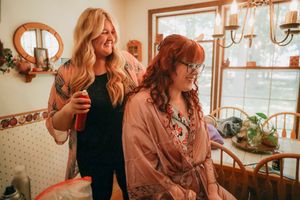 Hairstylist styling red-haired bride's hair in cozy home setting during wedding day preparations.