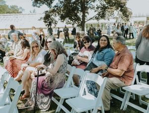 Guests seated in white chairs on grass at an outdoor wedding ceremony under a large tent on a sunny day.