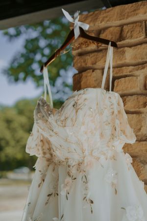 Elegant floral embroidered wedding dress hanging on a wooden hanger against a rustic stone wall outdoors.