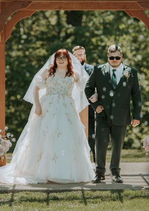Bride in white gown and groom in dark suit walk down aisle under wooden arch at outdoor wedding ceremony.