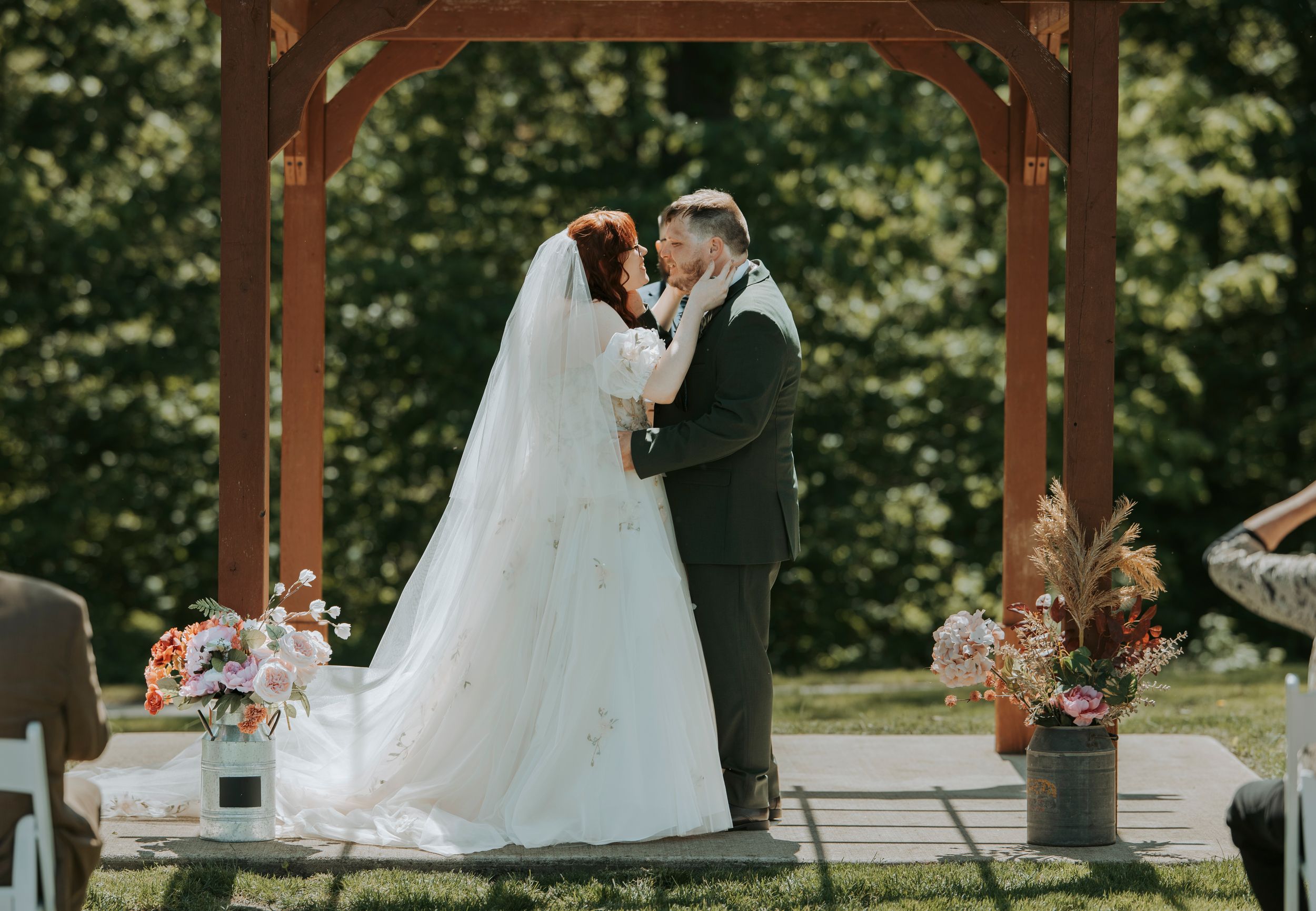 Bride and groom share first kiss under rustic wooden arbor at outdoor wedding ceremony with floral arrangements.