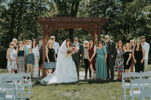 Wedding party poses with bride and groom under red pergola on sunny day outdoors.