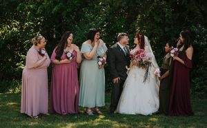 Wedding party poses outdoors with bride and groom surrounded by bridesmaids in pink and sage green gowns holding bouquets.