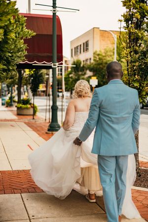 A couple in wedding attire walking together on a brick sidewalk past storefronts and lamp posts.