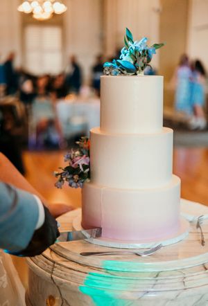 White wedding cake with turquoise accent lighting displayed on a rustic wooden platform at an indoor reception.