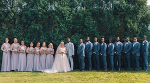 Wedding party members in coordinated attire stand in a row on a grassy hillside for a panoramic photo.