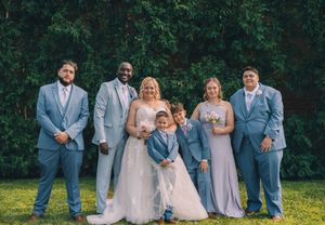 Wedding party in light blue and blush attire poses together on green lawn with forest backdrop.