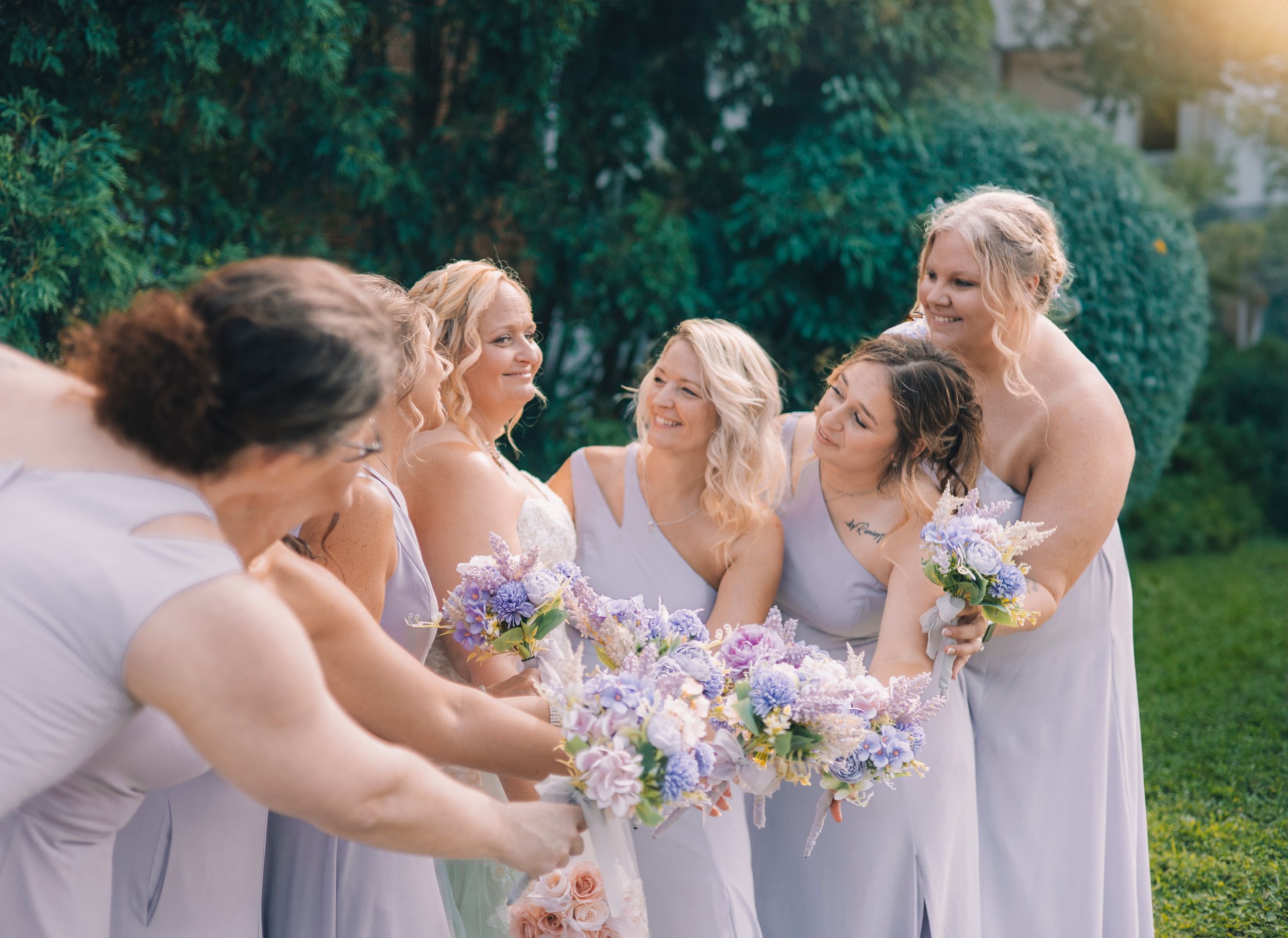 A group of bridesmaids in lavender dresses share a candid moment while holding purple and white bouquets outdoors.