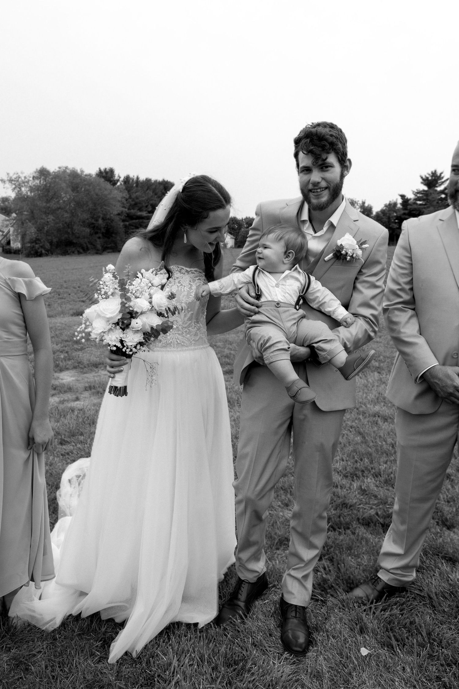 Wedding party shares a joyful moment with a baby outdoors in black and white.