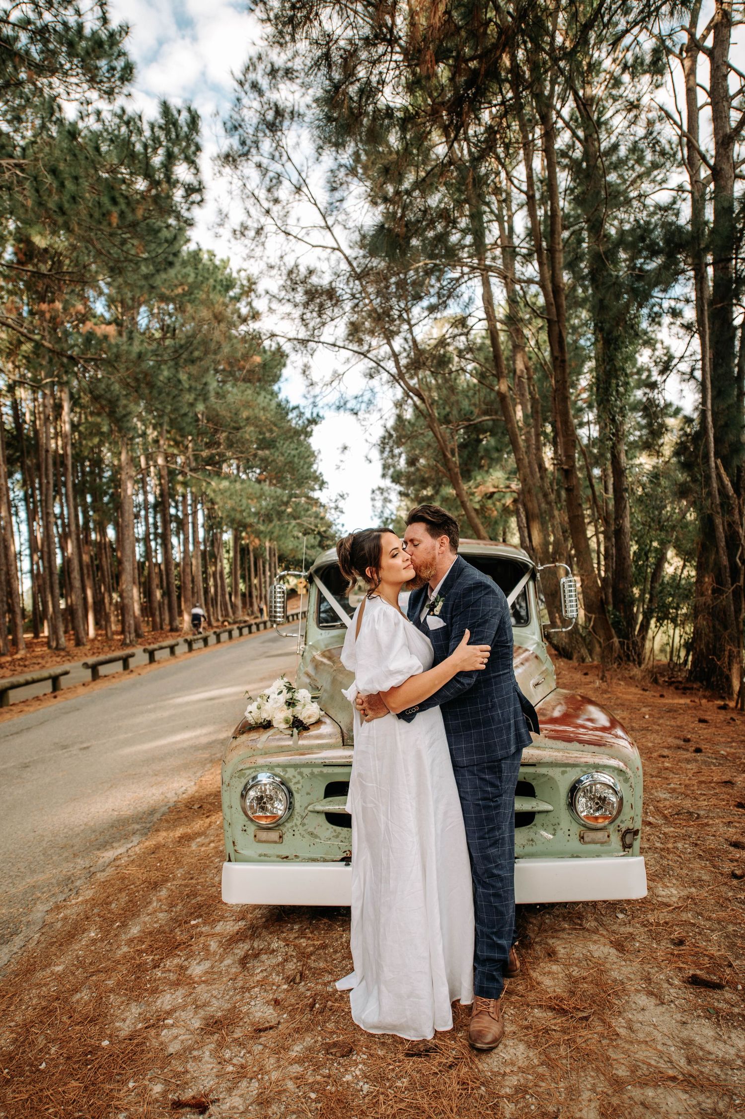 Bride and groom embrace against mint green vintage truck on forest path.