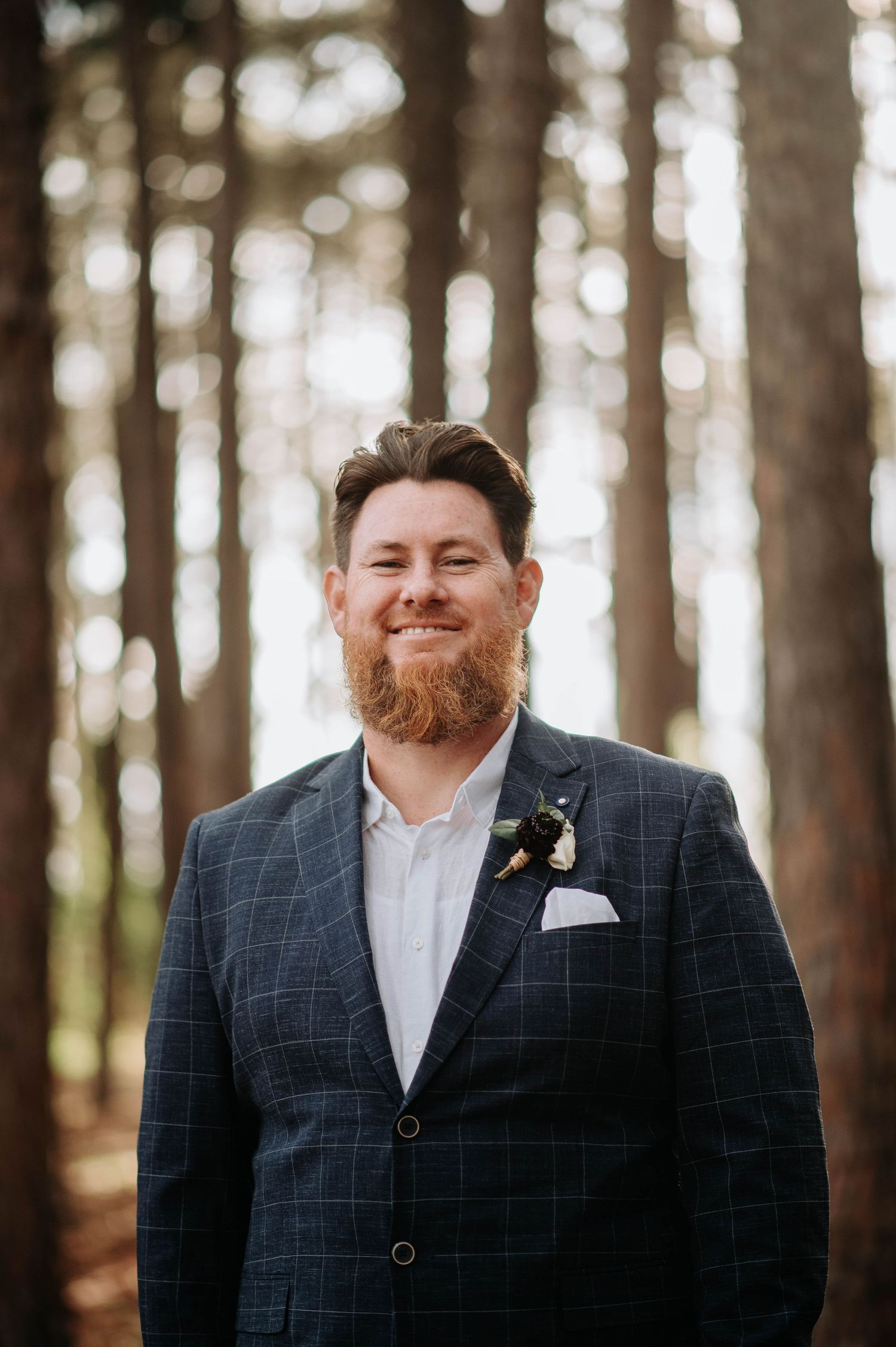 A groom in a checked navy suit with boutonniere stands in a sunlit forest.