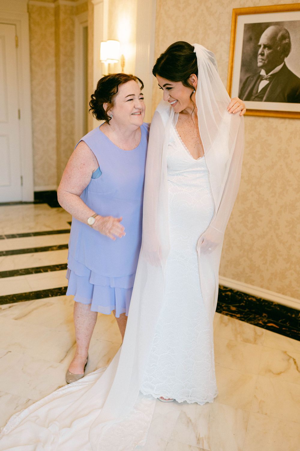 A bride in a white wedding gown shares a joyful moment with a woman in a light blue dress.