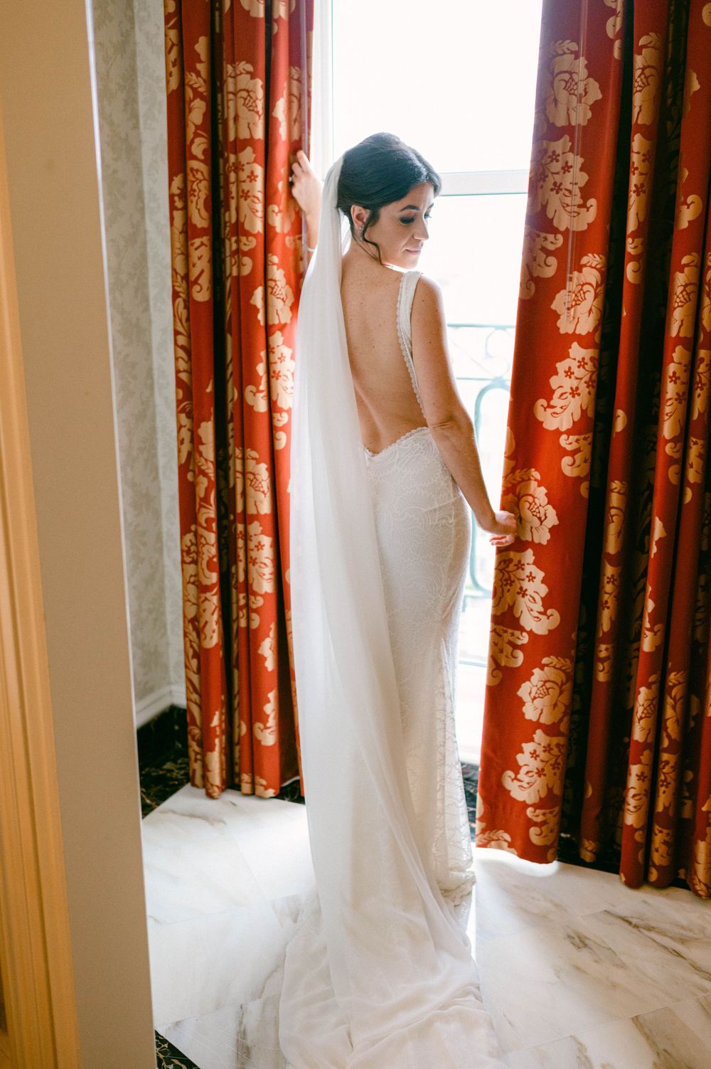 Elegant bride poses in a form-fitting backless wedding dress with long veil against red floral curtains.