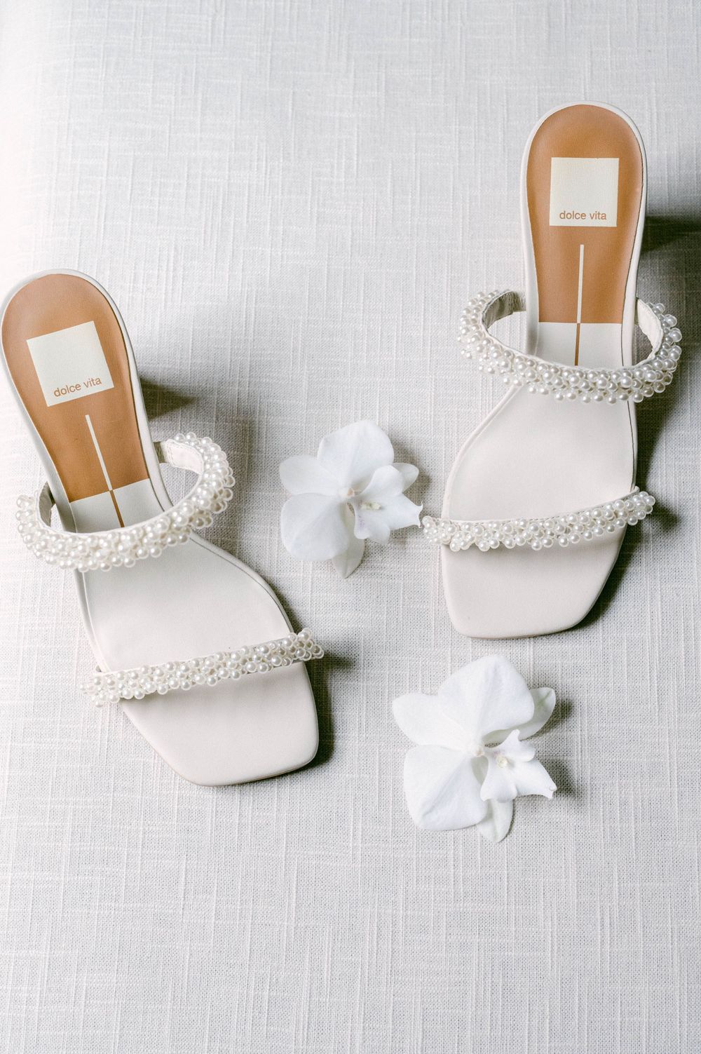 White bridal shoes with pearl embellishments displayed on light linen fabric.
