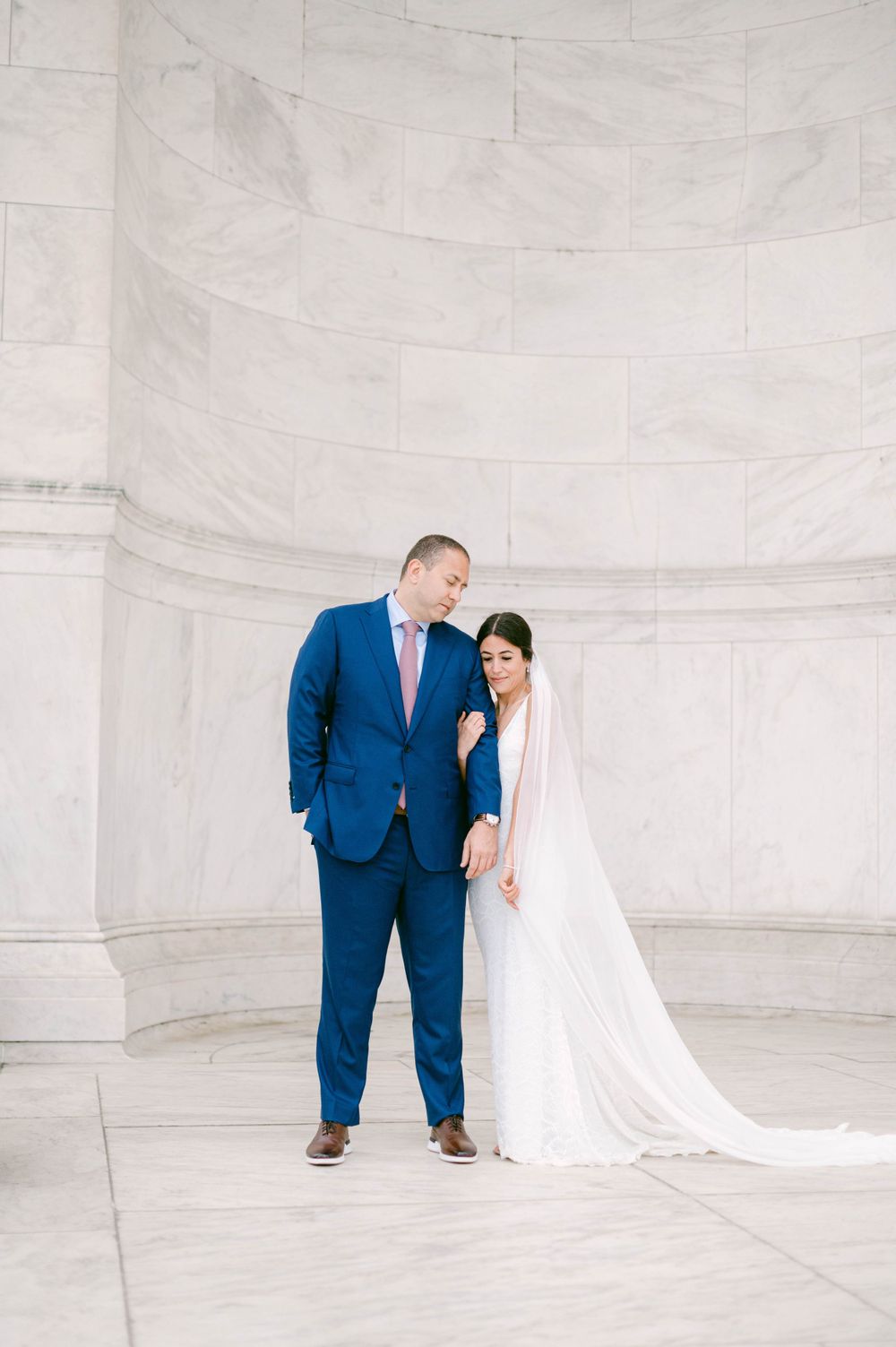 Wedding couple in blue suit and white dress posing against white marble backdrop.