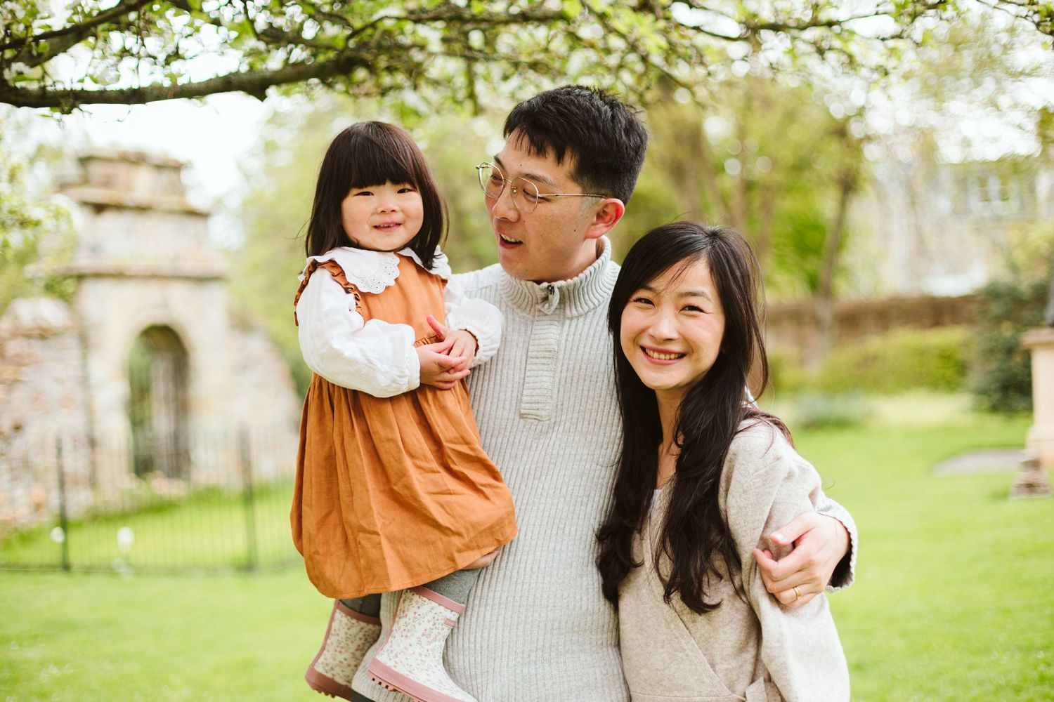 A happy family of three poses together on a grassy lawn with an old building in the background.