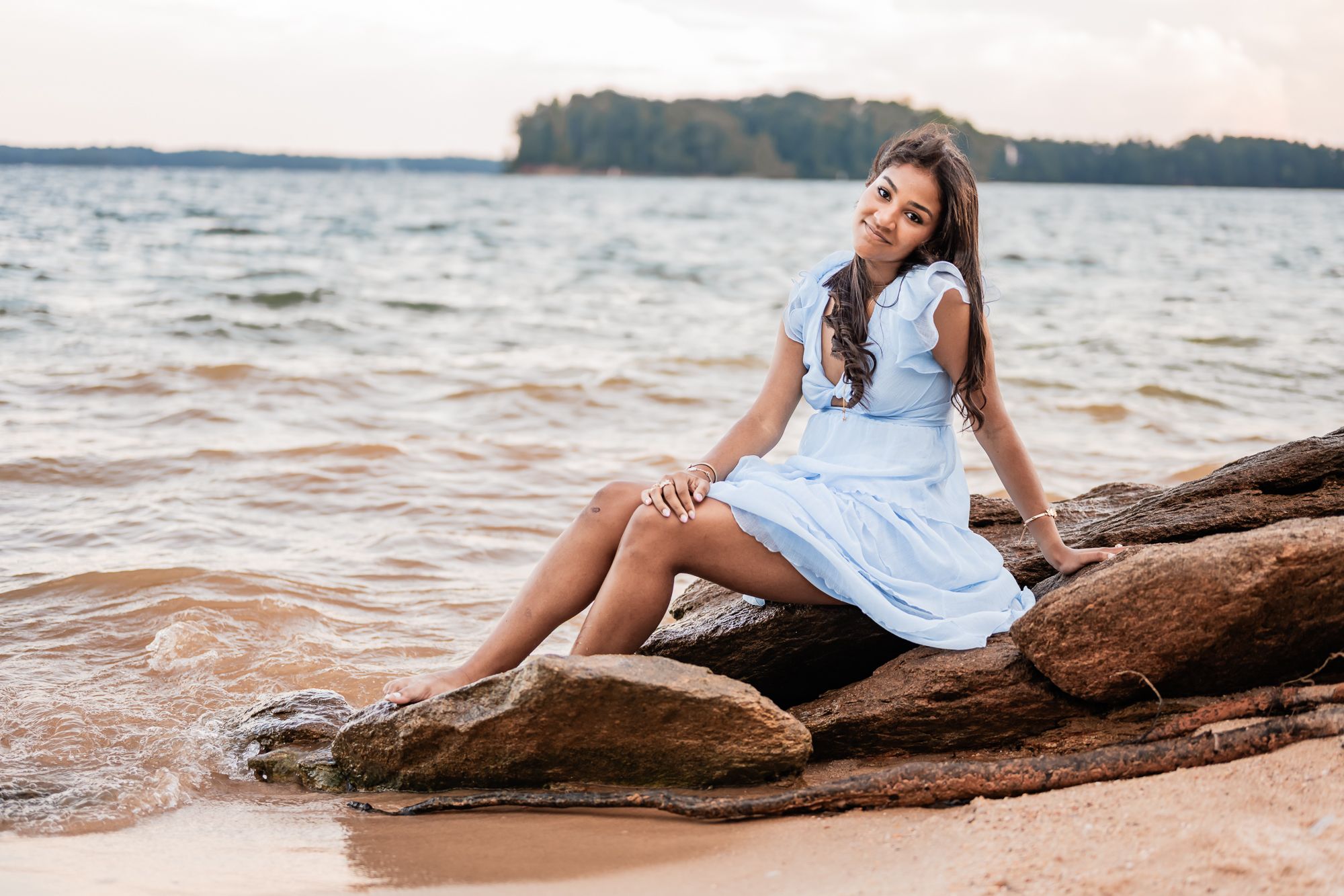 A person in a light blue dress sits gracefully on a driftwood log at the beach during sunset.
