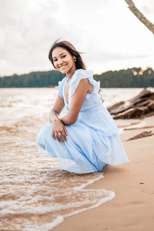 Woman in flowing light blue dress poses gracefully on sandy beach at sunset.