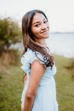 A person in a light blue ruffled dress poses outdoors against a natural blurred background.
