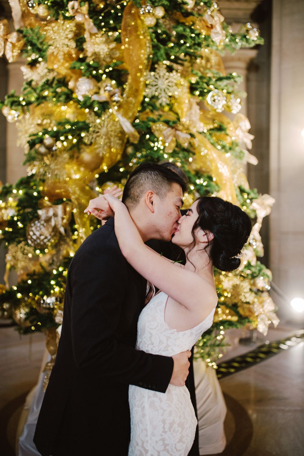 Couple shares romantic kiss in front of beautifully decorated Christmas tree with gold ornaments and lights.