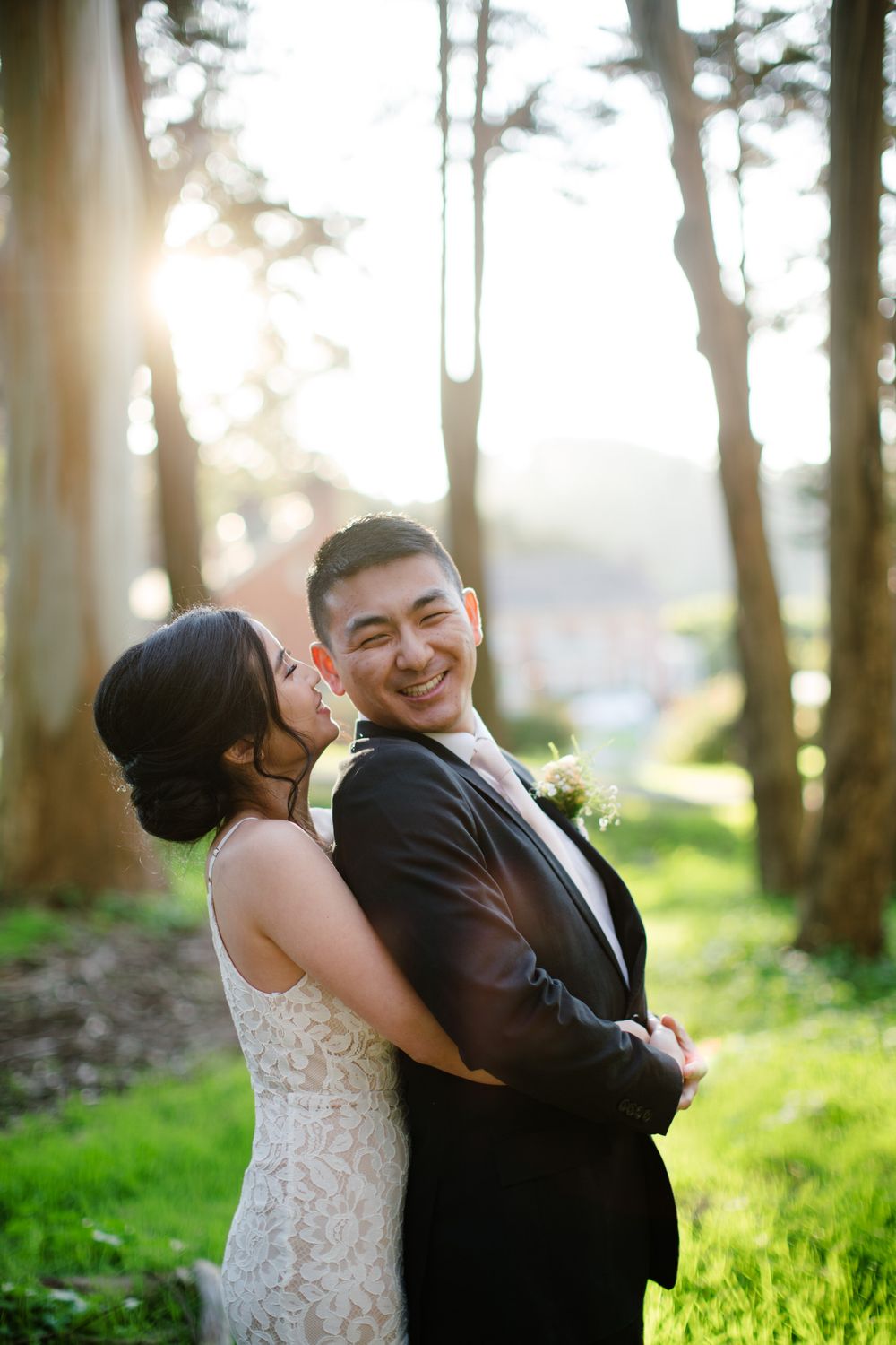 Couple in wedding attire laughing together in sunlit forest setting.