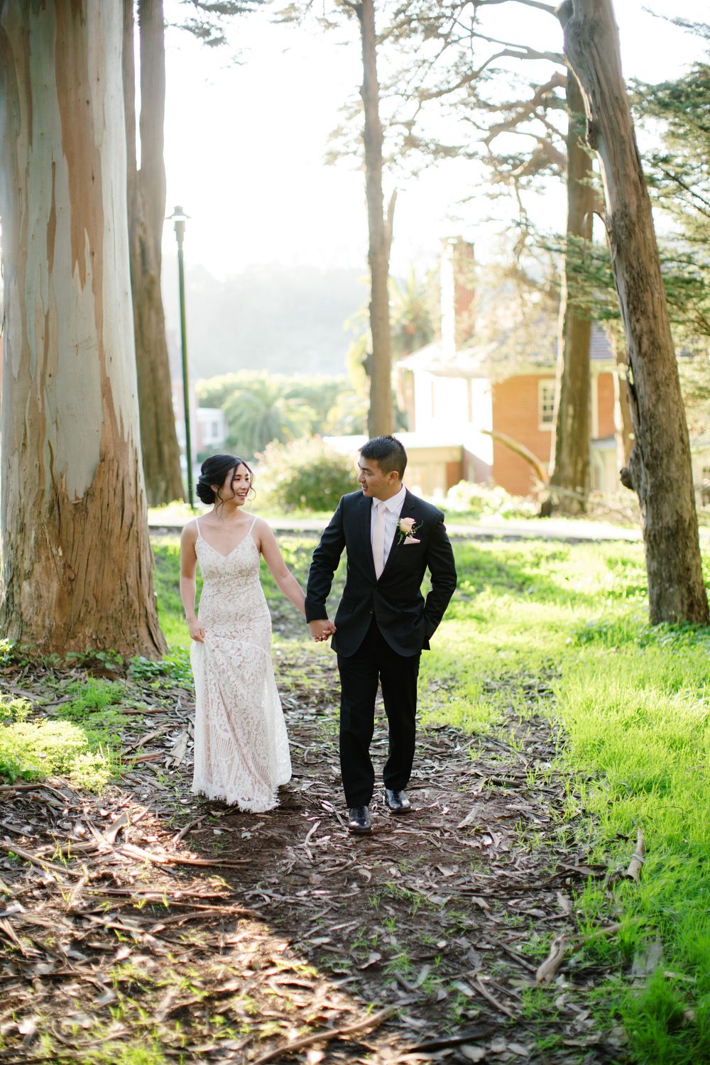 Bride in lace gown and groom walking through eucalyptus grove on wedding day.