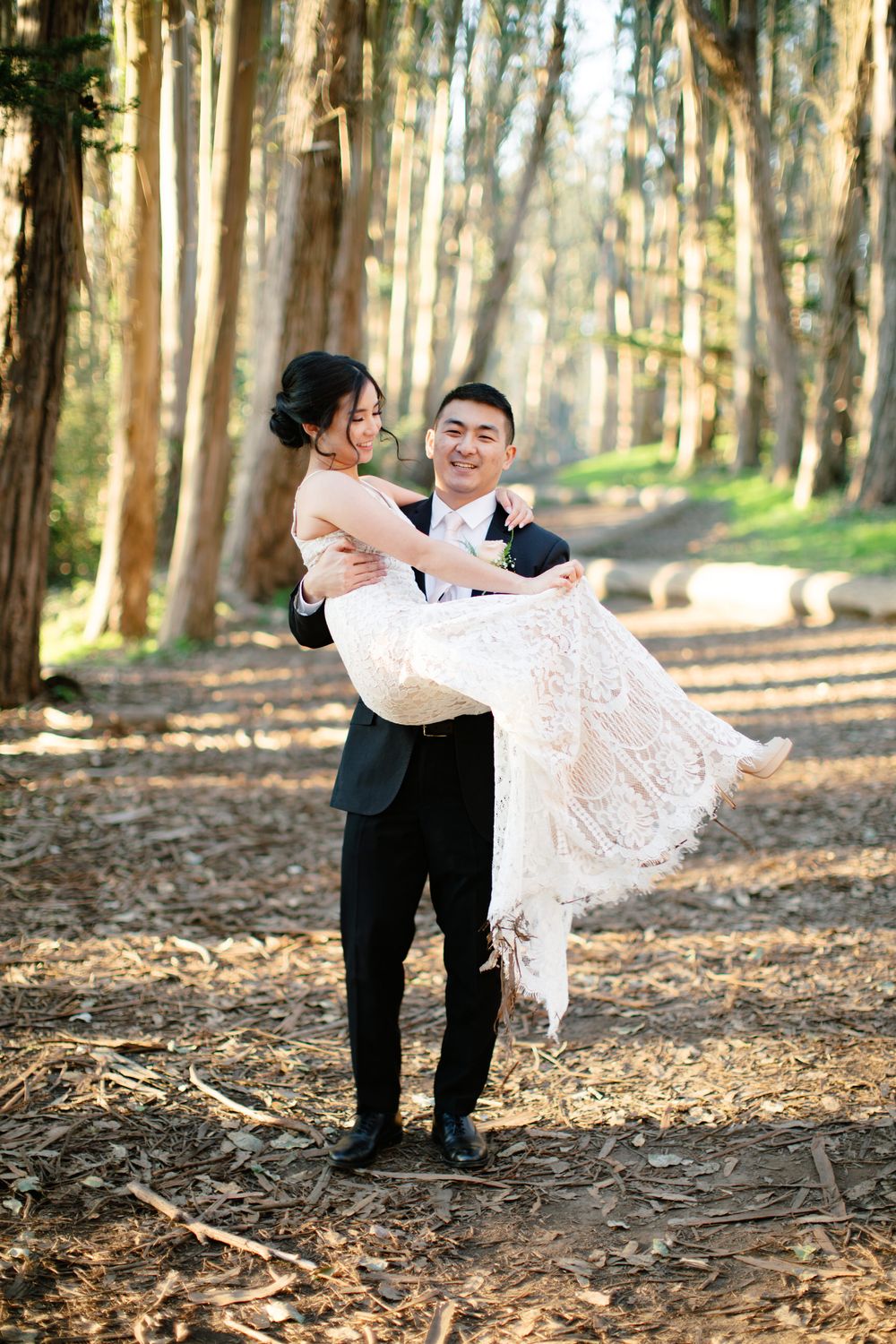 Groom lifts bride in lace wedding dress among tall trees in forest setting.