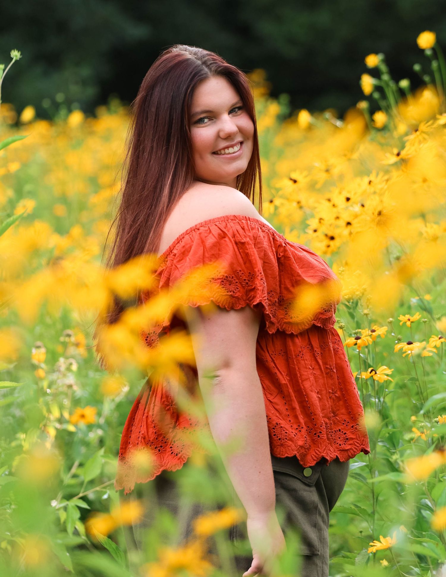 Portrait in a field of bright yellow wildflowers with a coral off-shoulder top against lush green foliage.