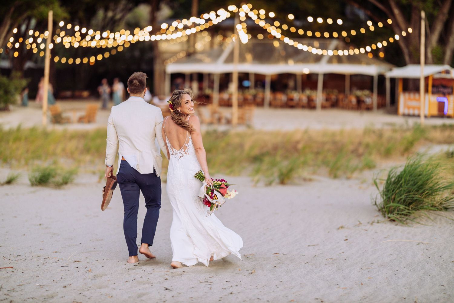 Couple walking on beach with string lights and wedding attire at dusk.