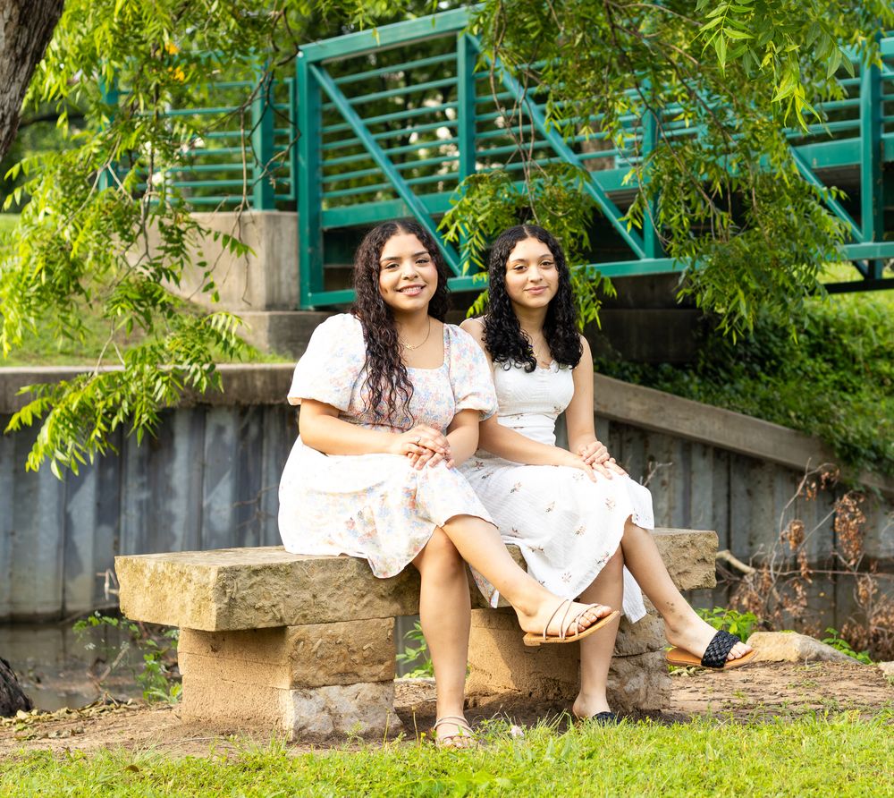 Two people in white dresses sit together on a stone bench near a turquoise structure surrounded by leafy trees.