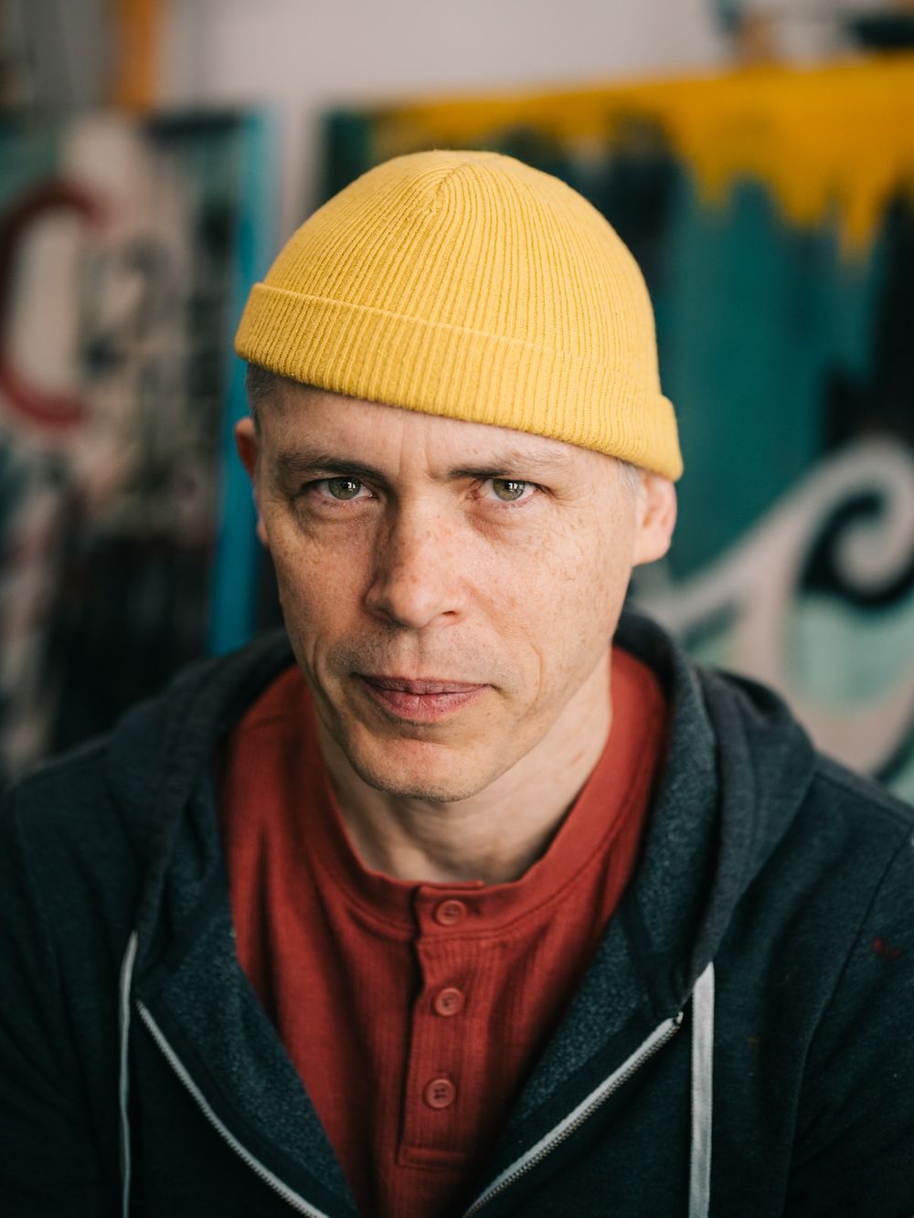 Person in yellow beanie and red henley shirt against colorful urban background.