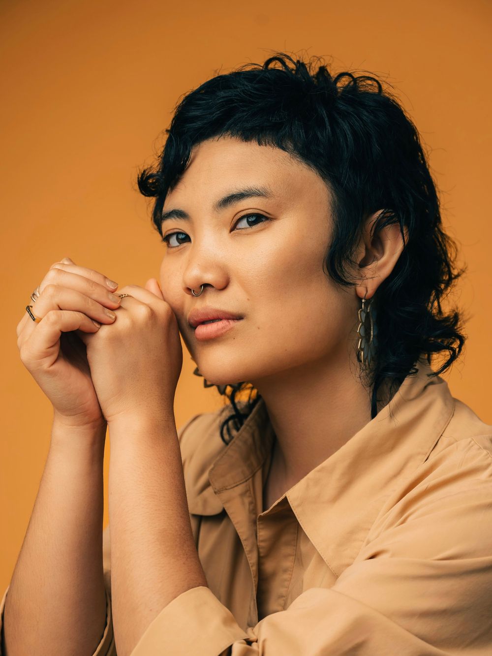 Professional portrait of subject with short dark curly hair wearing a beige button-up shirt against an orange background.