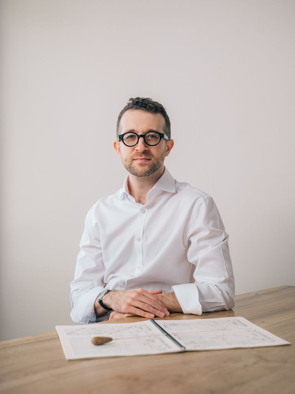 Professional wearing a white button-down shirt and glasses sits at wooden desk with documents and pen.