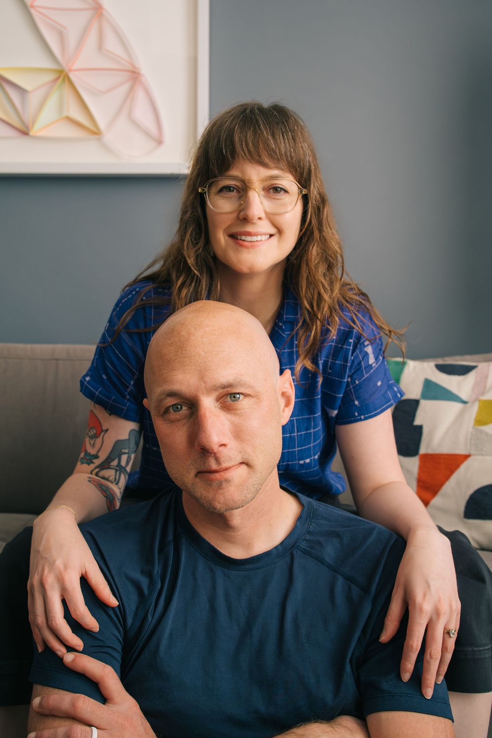 Couple sitting on grey couch at home with geometric wall art and throw pillows in background.