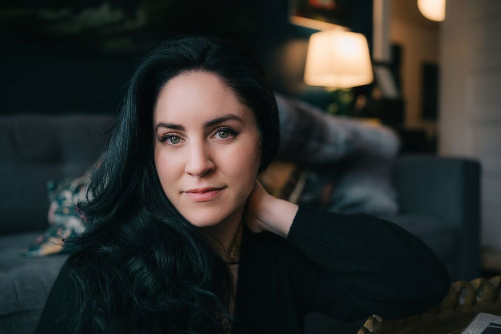 Indoor portrait of person with long dark hair resting hand on neck near grey couch.