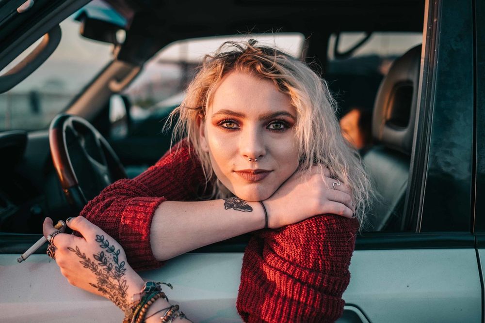 Portrait leaning on car door wearing red sweater with tattoos visible.