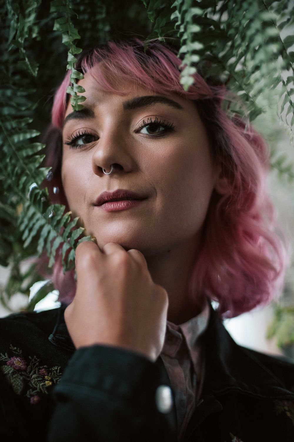 Portrait with pink hair framed by ferns in dramatic natural lighting.