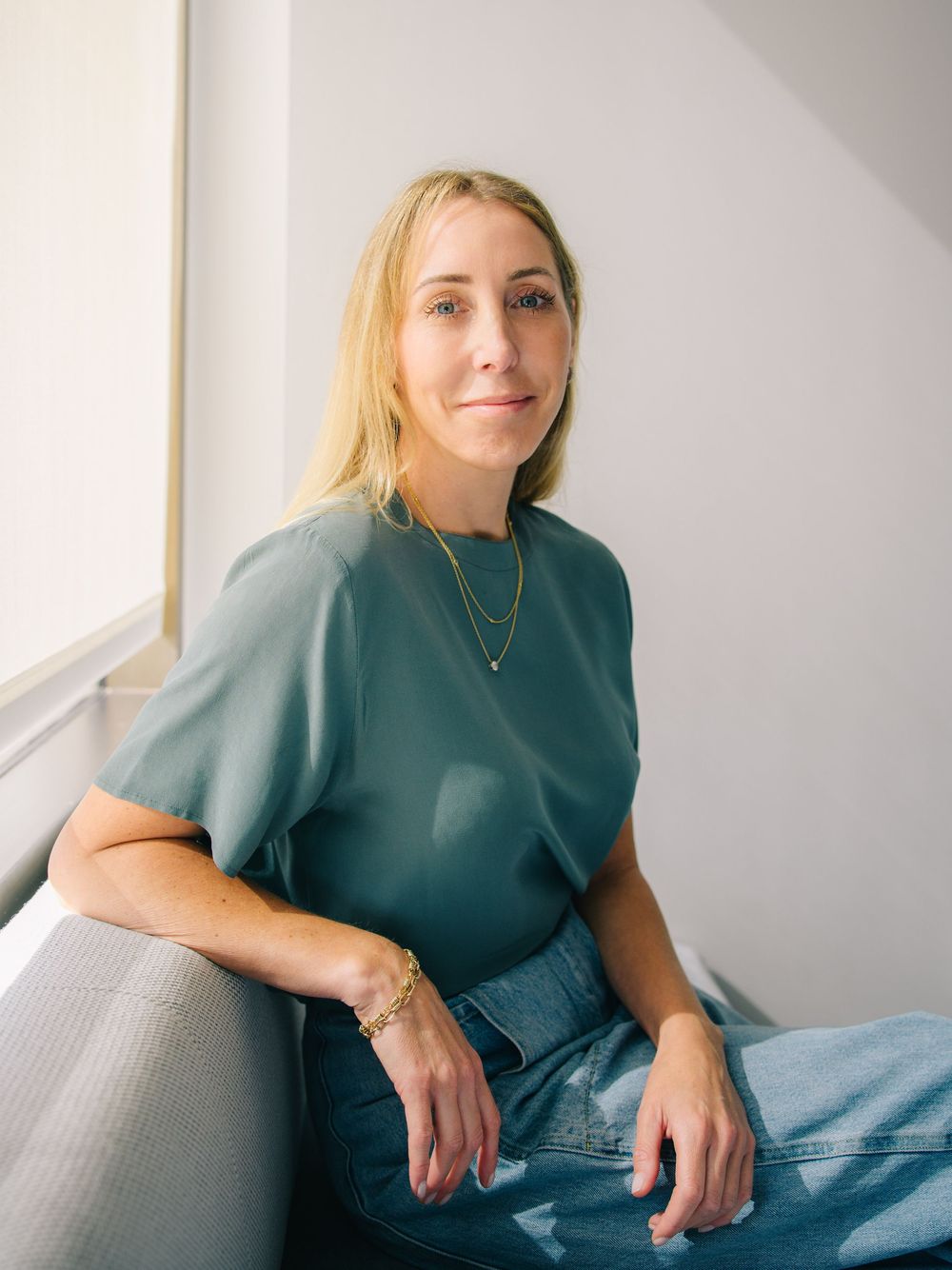 A person in a teal sweater and jeans sits casually by a window in natural light.