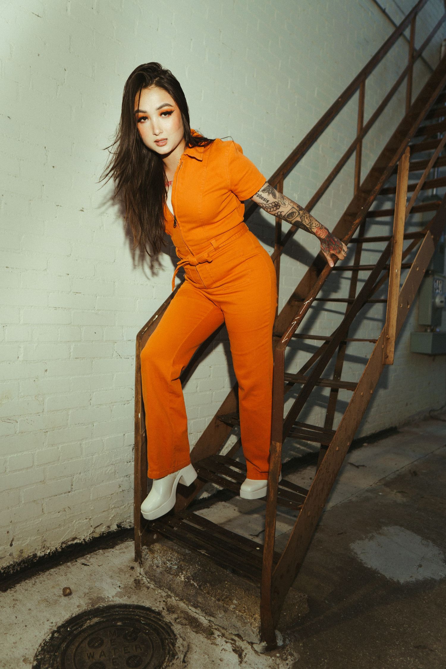 A model poses on metal stairs wearing a vibrant orange jumpsuit and white boots in an industrial setting.
