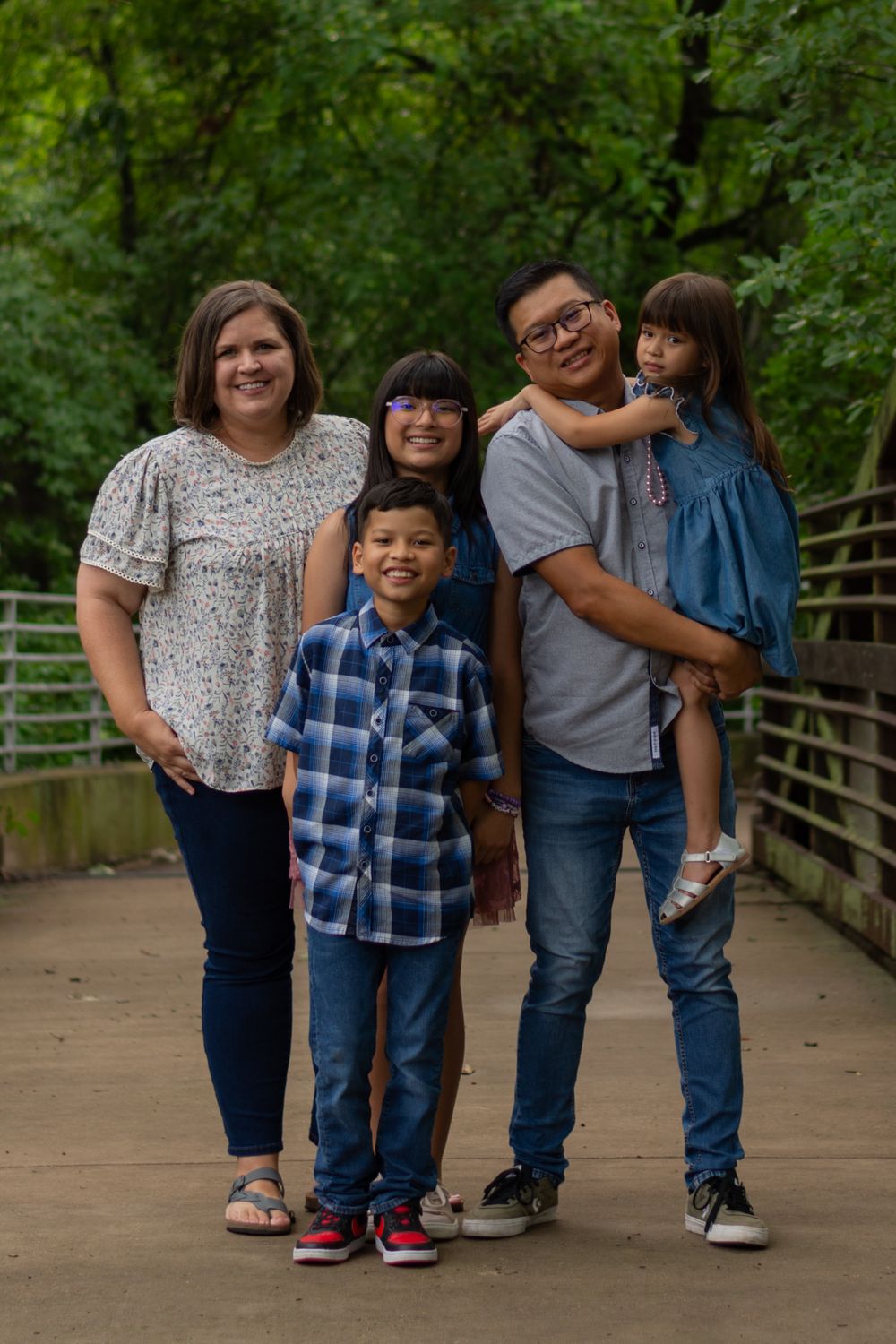A family poses together on a bridge at a Fort Worth park on an overcast, cloudy day. Dad is holding the youngest child in his arms.