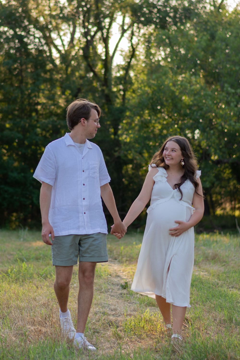 A couple walks in nature, looking at each other lovingly, during a summer maternity photo session in a Fort Worth park. She holds her belly.