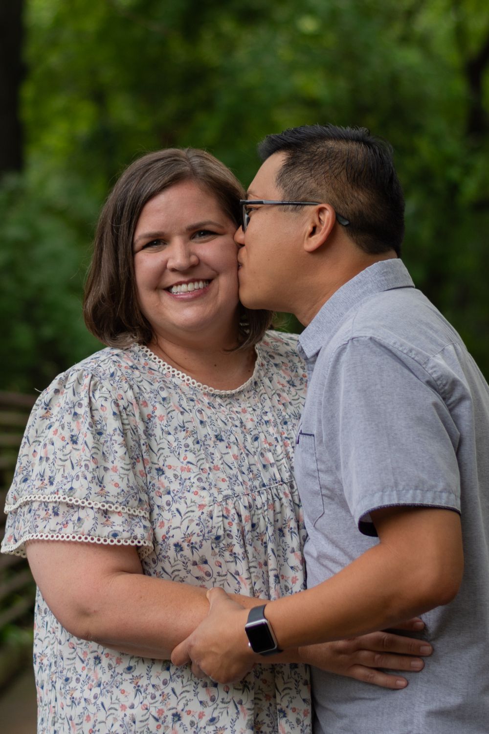 A husband kisses his wife's cheek as she smiles at the camera during a family photoshoot in a park in Fort Worth.