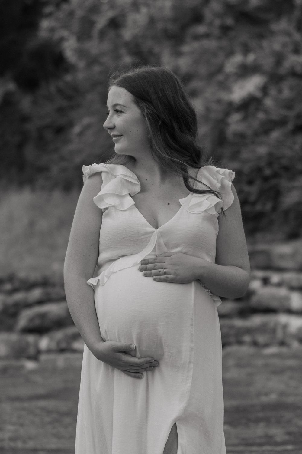 B&W photo of a pregnant mother-to-be looking off to the side as she holds her baby bump with both hands. She stands in a Fort Worth river.