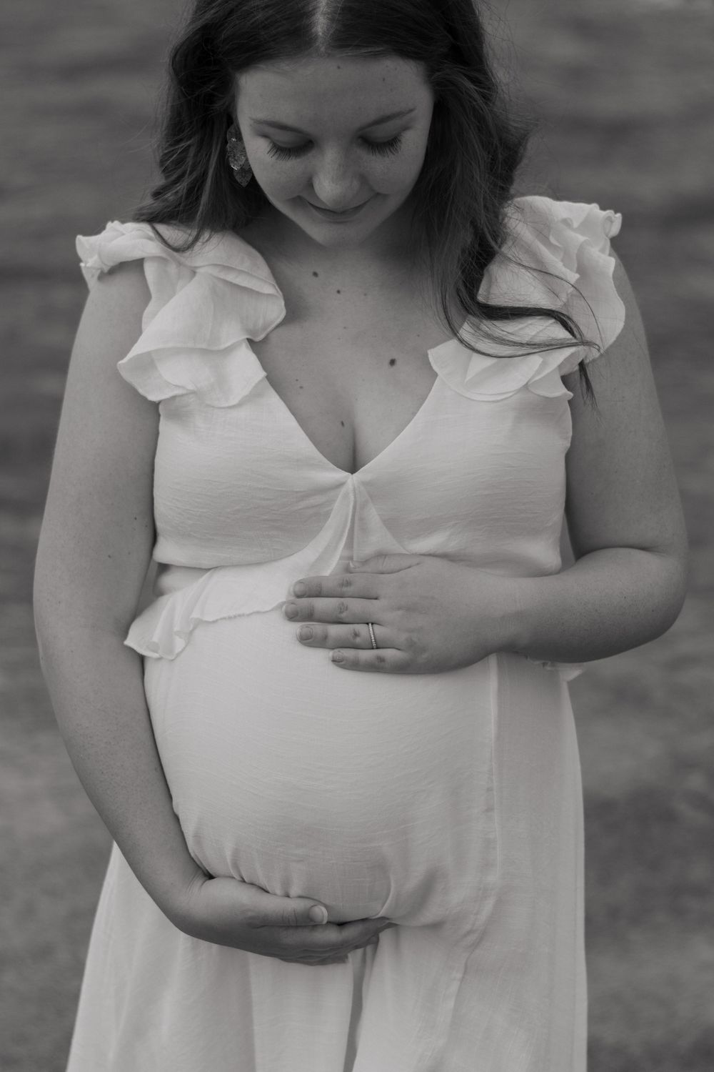 Black-and-white of a pregnant woman standing in a river in her white dress, looking down while holding her baby bump with both hands.