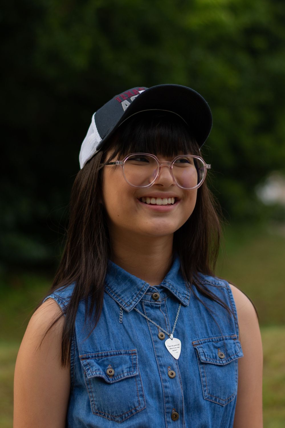 A teen girl poses for a photo during a family session while wearing her dad's Aggie hat. Photoshoot located in Fort Worth, Texas.