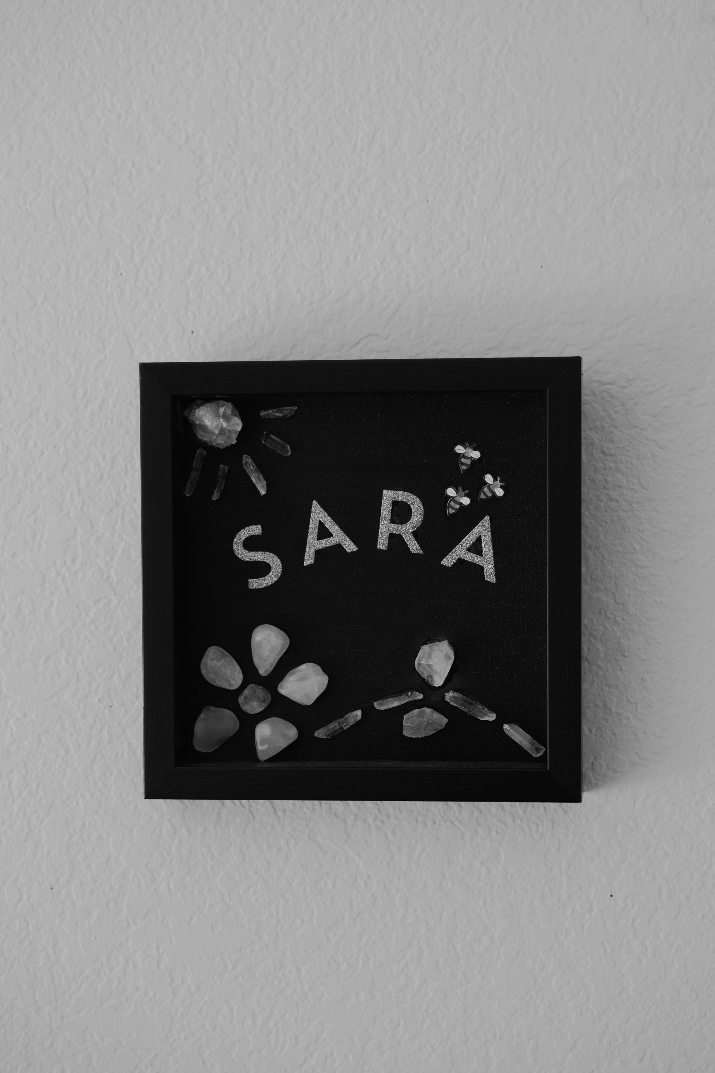 Black framed artwork displaying 'SARA' text with floral decorative elements.
