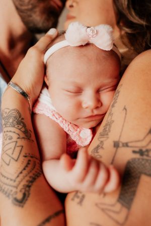 Sleeping newborn with white bow headband held by parents with visible tattoos.